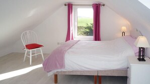 A bedroom with a bed, a chair, a nightstand, a lamp, and a window with curtains. - The White Cottage Leap (Reenascreena)