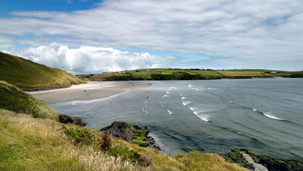 A coastal landscape with a sandy beach, grassy hills, and a body of water. - The White Cottage Leap (Reenascreena)