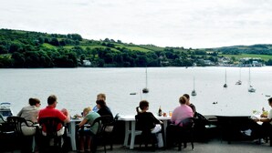 A lakeside restaurant with tables and chairs facing the water, where people are dining. - The White Cottage Leap (Reenascreena)