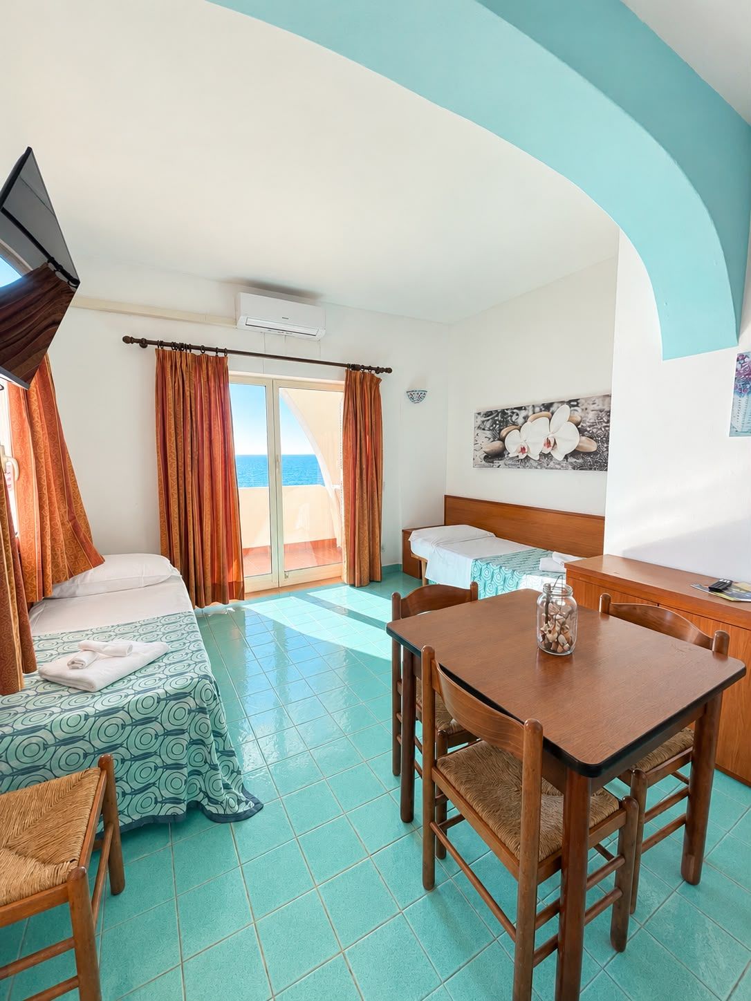 A hotel room with a bed, a desk, and a chair. There is a window with curtains, a flat-screen TV mounted on the wall, and a small table with a jar on it.