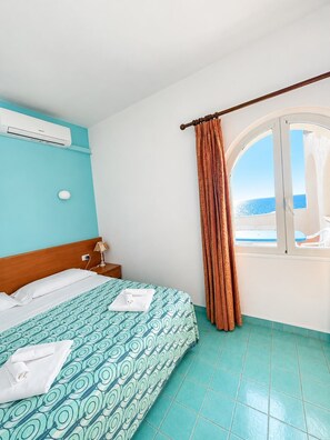 A hotel room with a bed, an air conditioning unit, a nightstand, a lamp, and a window with curtains. - Two-bedroom apartment, large terrace, ocean view, 1st floor (Pink) (Anzio)