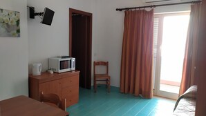 Habitación con kitchenette, una mesa de comedor, una silla, un armario de madera, microondas, hervidor de agua, televisor, una puerta y una ventana con cortinas.