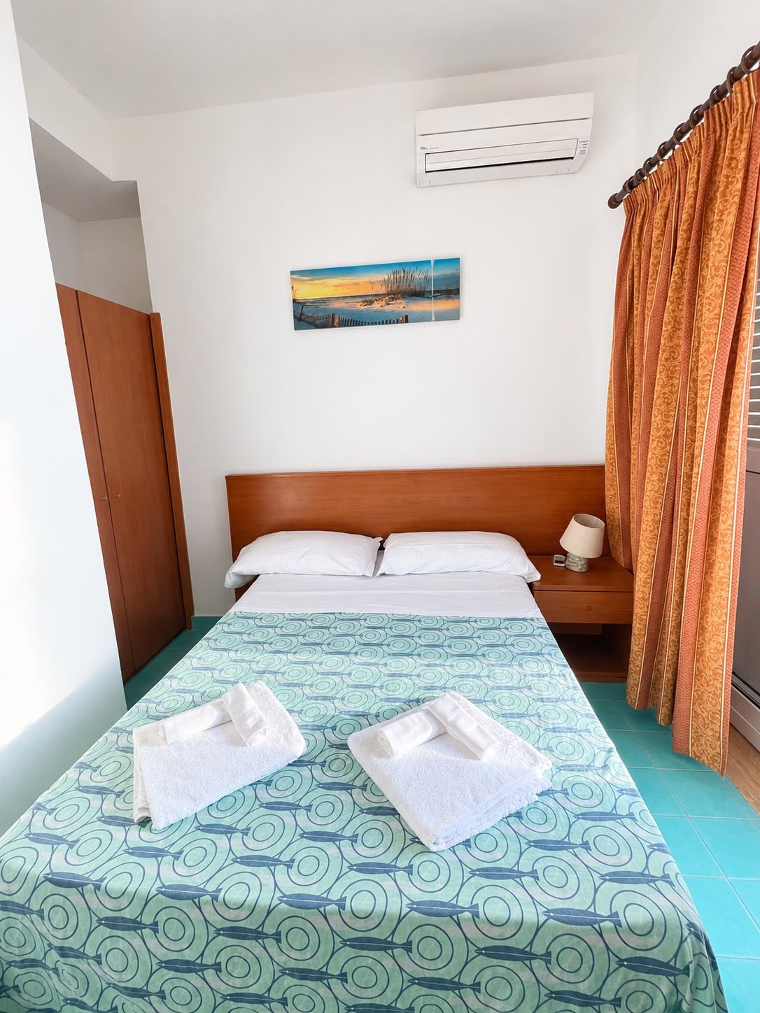 Een hotelkamer met een bed, nachtkastje, airconditioning, schilderij en gordijn.