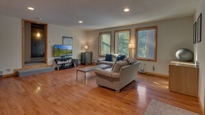 A spacious living room with hardwood flooring, a sofa, a coffee table, and a television. There are large windows with views of trees and a door leading to another room. - Chamberlands Cabin: 5  BR, 3  BA House in Tahoma, Sleeps 10 (Tahoma)