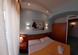 Une chambre d’hôtel comprenant un lit, une table de chevet, une lampe fixée au mur, un tableau et une armoire.