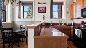 A kitchen with a central island, wooden cabinets, a dining table with chairs, and a window with a view of trees. - Papa's Place — Historic 1BR Brick Unit, Walk to Main St Leadville (Leadville)