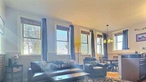 A living room with a sofa, armchairs, a coffee table, and a patterned rug. There are large windows with curtains, a lamp, and a dining area with chairs and a table. - Papa's Place — Historic 1BR Brick Unit, Walk to Main St Leadville (Leadville)