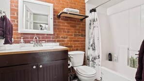 A bathroom with a brick wall, a wooden vanity, a white toilet, and a shower curtain. - Papa's Place — Historic 1BR Brick Unit, Walk to Main St Leadville (Leadville)
