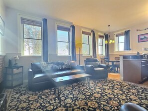 A living room with a sofa, armchairs, a coffee table, and a patterned rug. There are large windows with curtains, a lamp, and a dining area with chairs and a table.