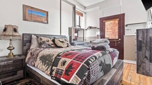 A bedroom with a bed, a nightstand, a lamp, a mirror, a door, and a window. - Papa's Place — Historic 1BR Brick Unit, Walk to Main St Leadville (Leadville)