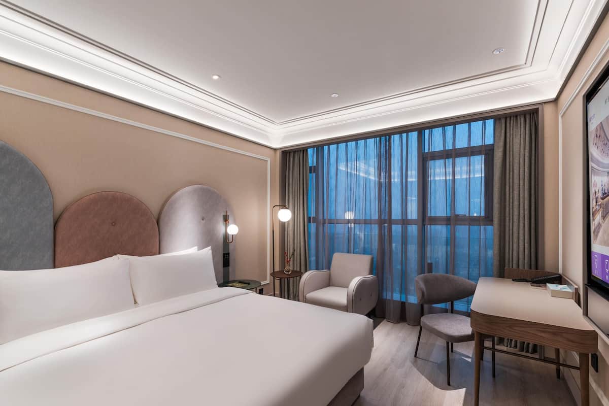A hotel room with a large bed, a desk, two chairs, a TV, and a large window with curtains.