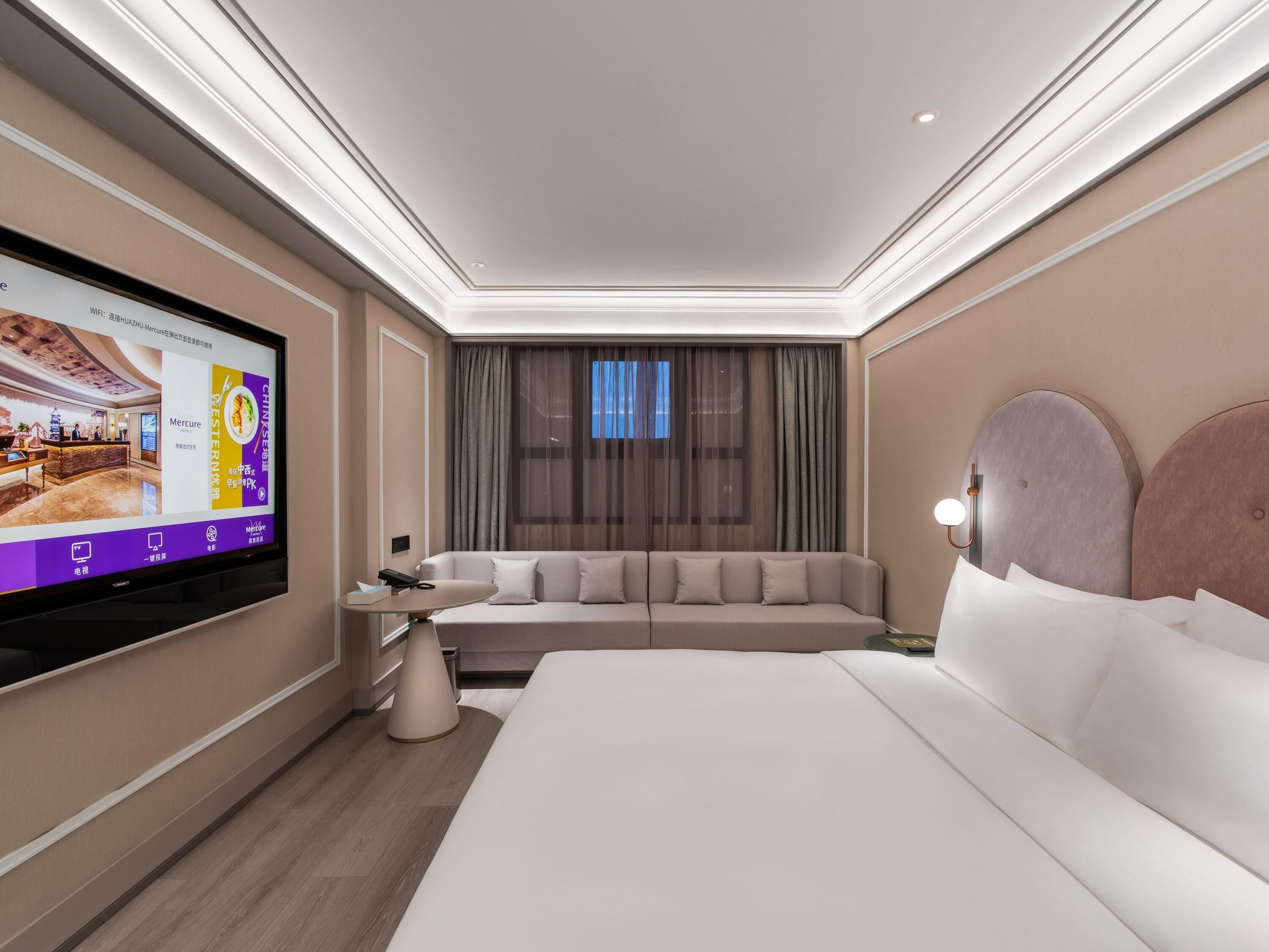 A modern hotel room with a large bed, a sofa, a TV on the wall, and a window with curtains.