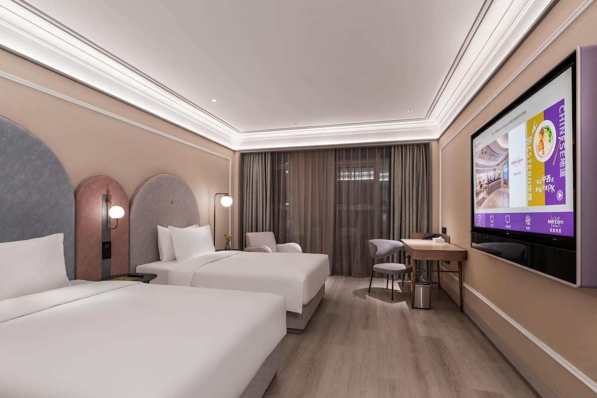 A hotel room with two beds, a desk, a chair, a TV screen, and a large window with curtains.