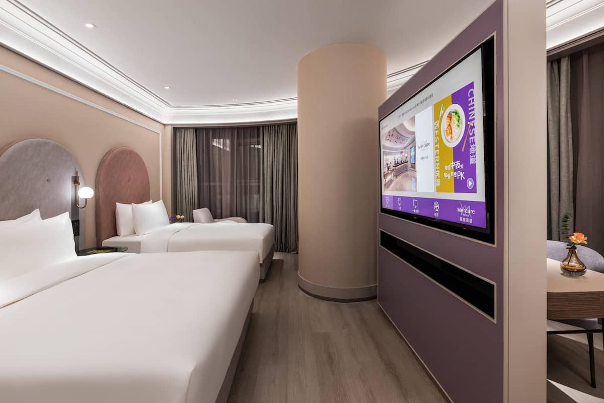 A hotel room with two beds, a TV mounted on the wall, and a large window with curtains.