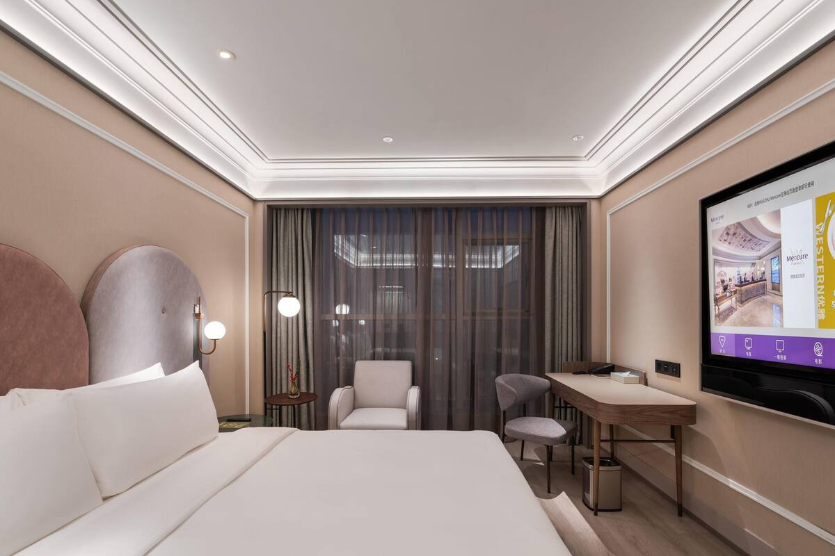 A modern hotel room with a large bed, a desk, a chair, a TV, and a large window with curtains.