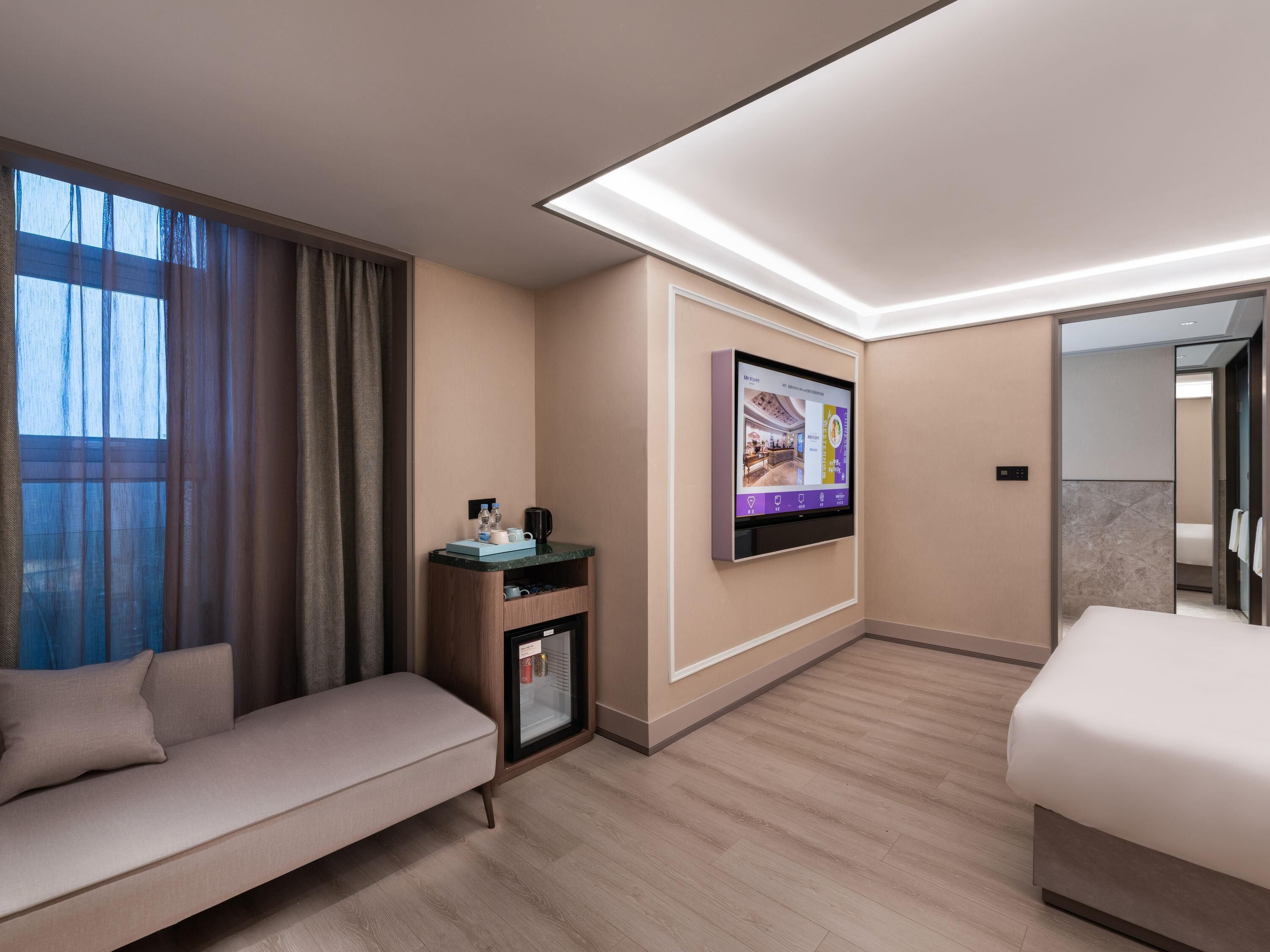 A hotel room with a sofa, a TV mounted on the wall, a small table with a coffee maker, and a minibar.