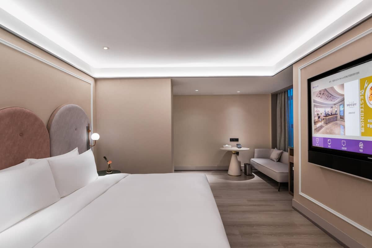 A modern hotel room with a large bed, a small table, a sofa, and a TV displaying a hotel interior.