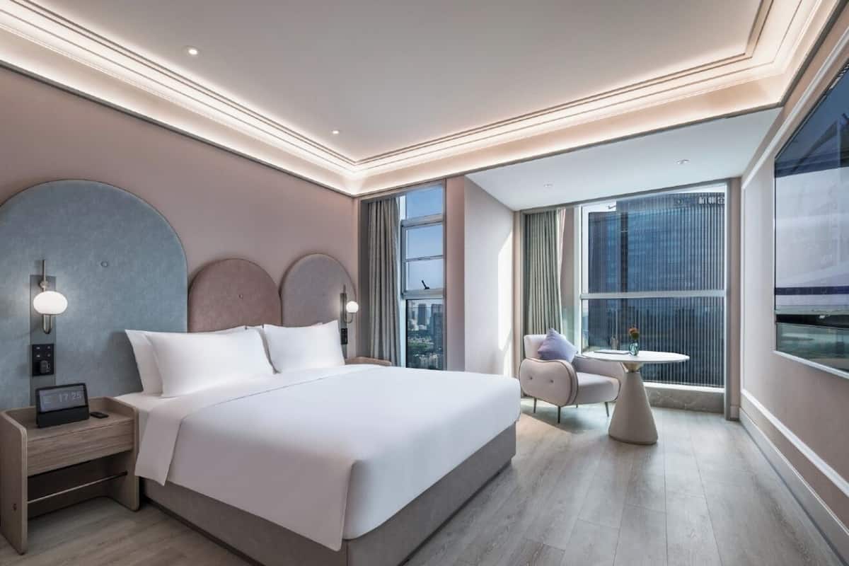A modern hotel room with a large bed, a bedside table with a clock, a small round table with a chair, and a large window offering a city view.