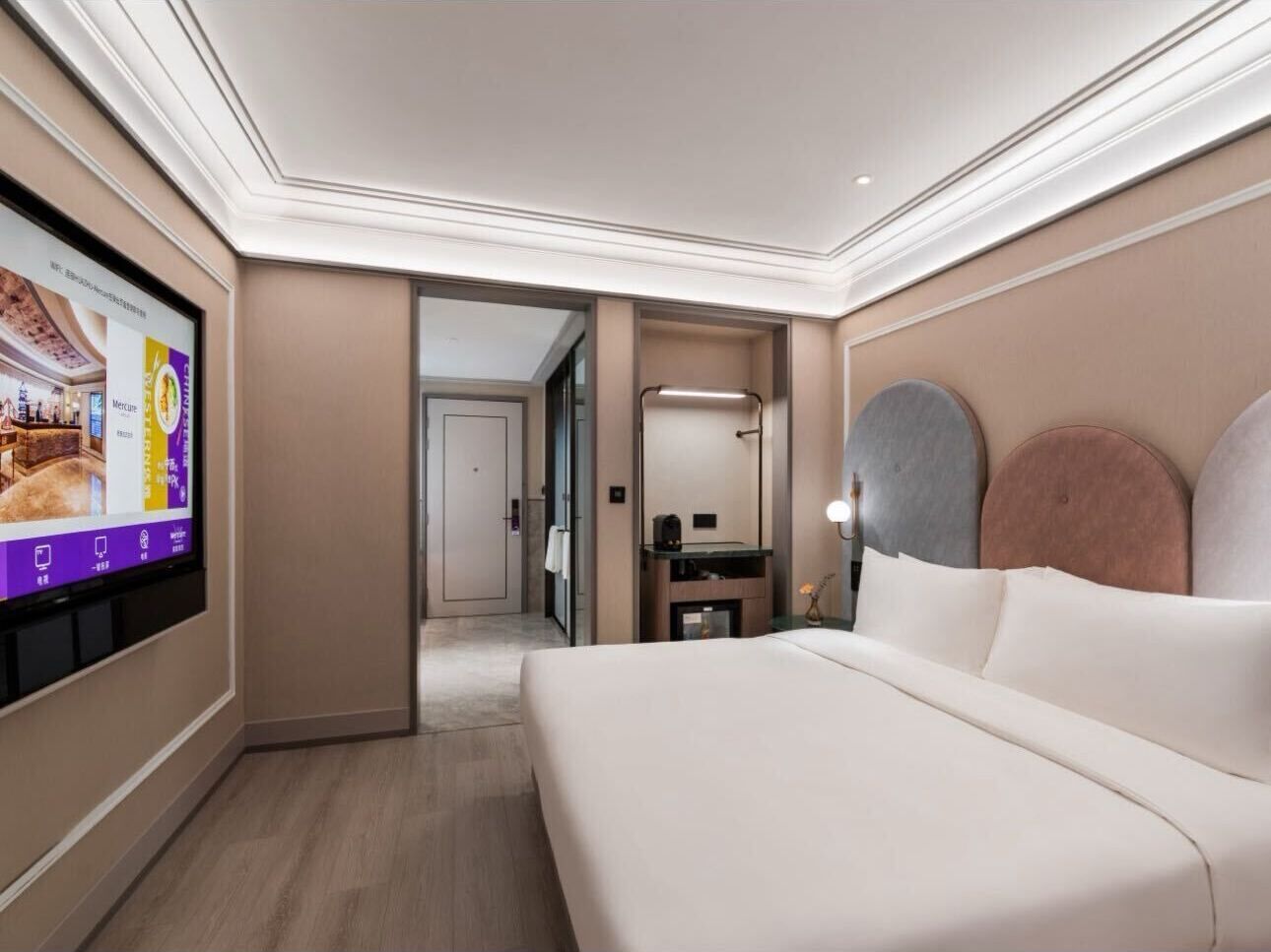 A hotel room with a large bed, a TV on the wall, a desk with a mirror, and a door leading to another room.