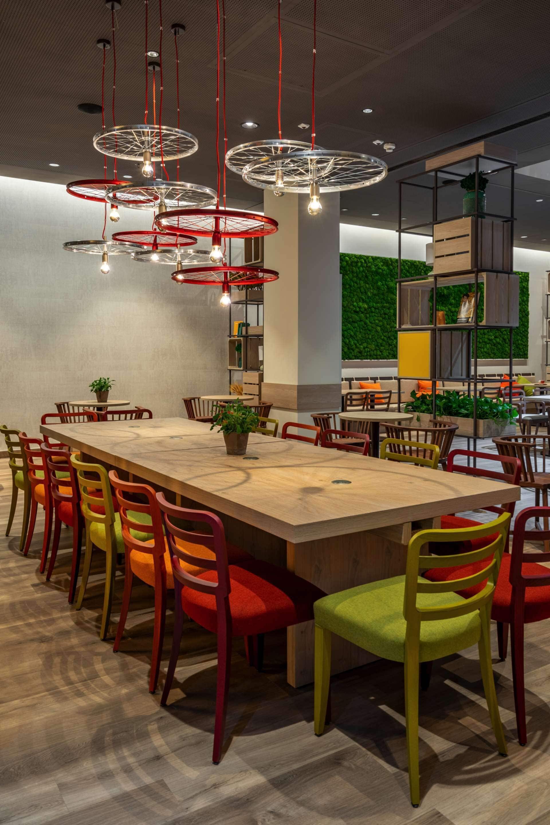 A modern dining area with a long wooden table, colorful chairs, and hanging red light fixtures.