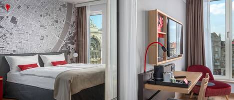 A modern hotel room with a large bed, a desk with a chair, a mirror, and a view of the cityscape.