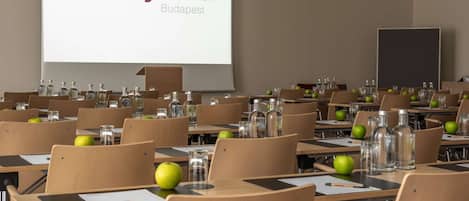 Un salón de conferencias con filas de escritorios, sillas y una pantalla de proyector que muestra