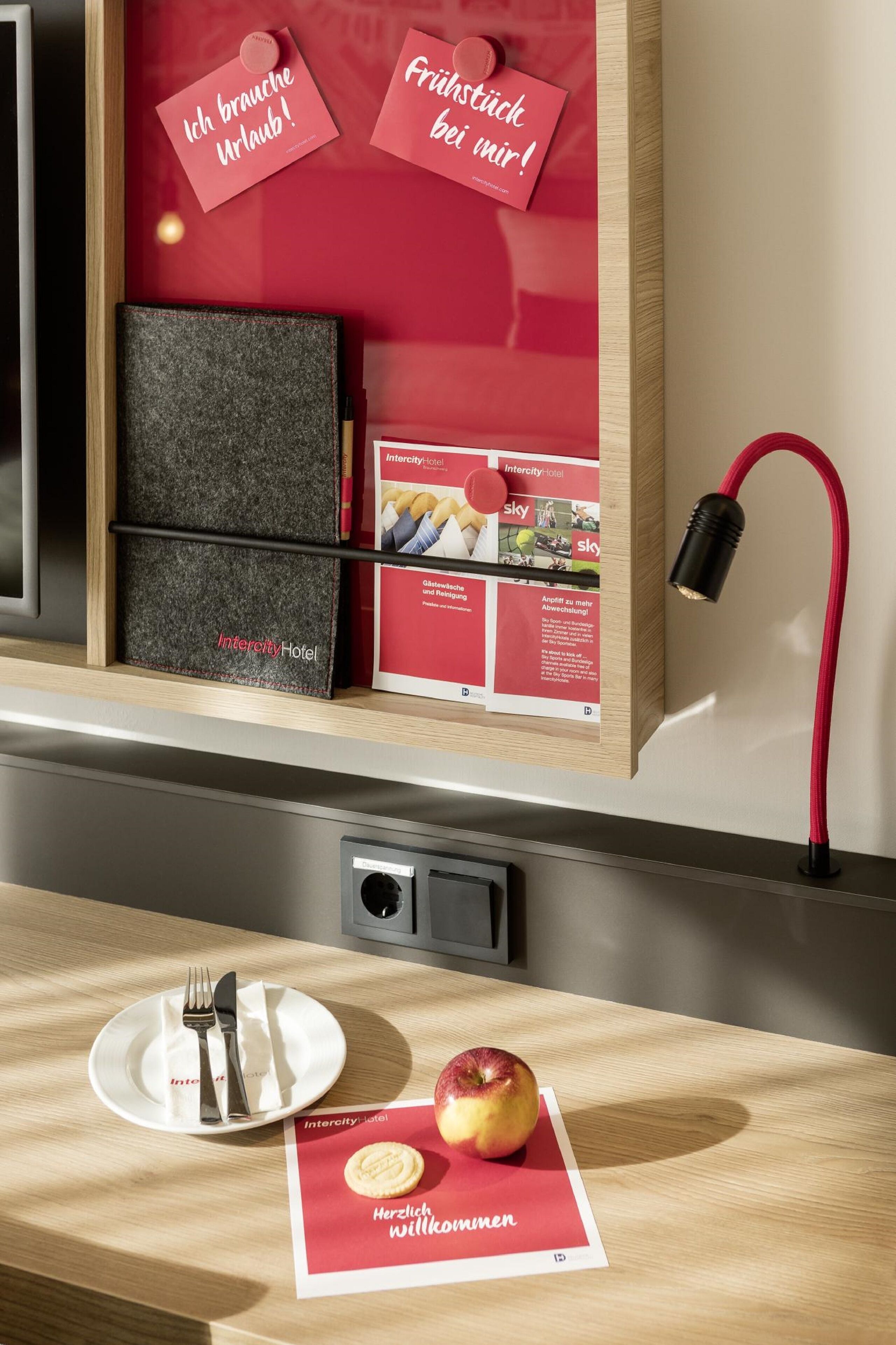 A hotel room with a wooden desk, a plate with cutlery, an apple, and a welcome card.