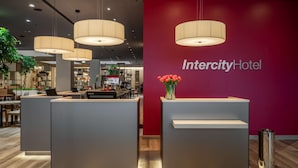 A reception area with a red wall, a Lenovo desk, and a vase of red flowers. - IntercityHotel Budapest (Budapest)
