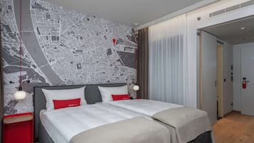 Quarto de hotel com um mapa grande na parede, uma cama com roupa de cama branca e almofadas vermelhas, uma mesa de cabeceira vermelha pequena e piso de madeira.