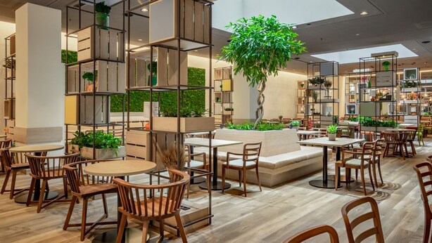 A modern indoor space with wooden tables and chairs, a central plant, and built-in shelving units. - IntercityHotel Budapest (Budapest)