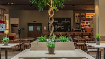 Um espaço interior moderno com uma mesa de madeira central rodeada por cadeiras, uma grande planta em vaso e uma área de estar com bancos acolchoados.