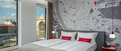 A modern hotel room with a large bed, a cityscape view, a map wall, and a red accent wall.