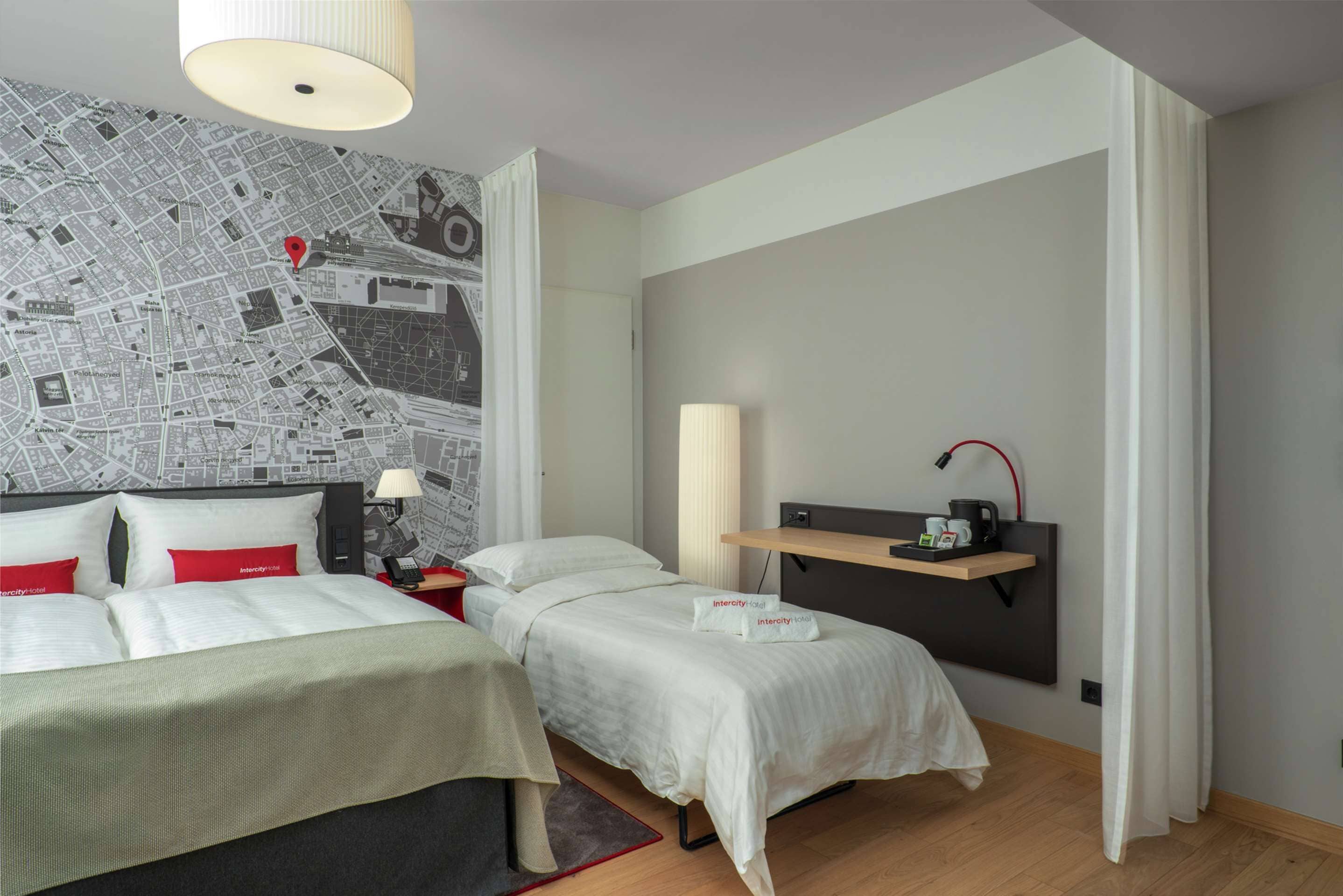 Una habitación de hotel con un mapa grande en la pared, una cama con ropa de cama blanca, una mesita de noche con una lámpara y un estante con cafetera.