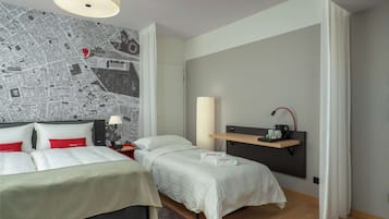 Une chambre d’hôtel avec une grande carte accrochée au mur, un lit recouvert de draps blancs, une table de chevet avec une lampe et une étagère sur laquelle se trouve une machine à café.