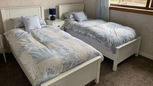Quarto com duas camas de solteiro, mesa de cabeceira com abajur, relógio na parede e janela com vista para um campo verde.