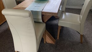 Uma área de refeições com uma mesa de madeira, cadeiras brancas e uma tigela verde. Há uma janela com cortinas e um radiador na parede.