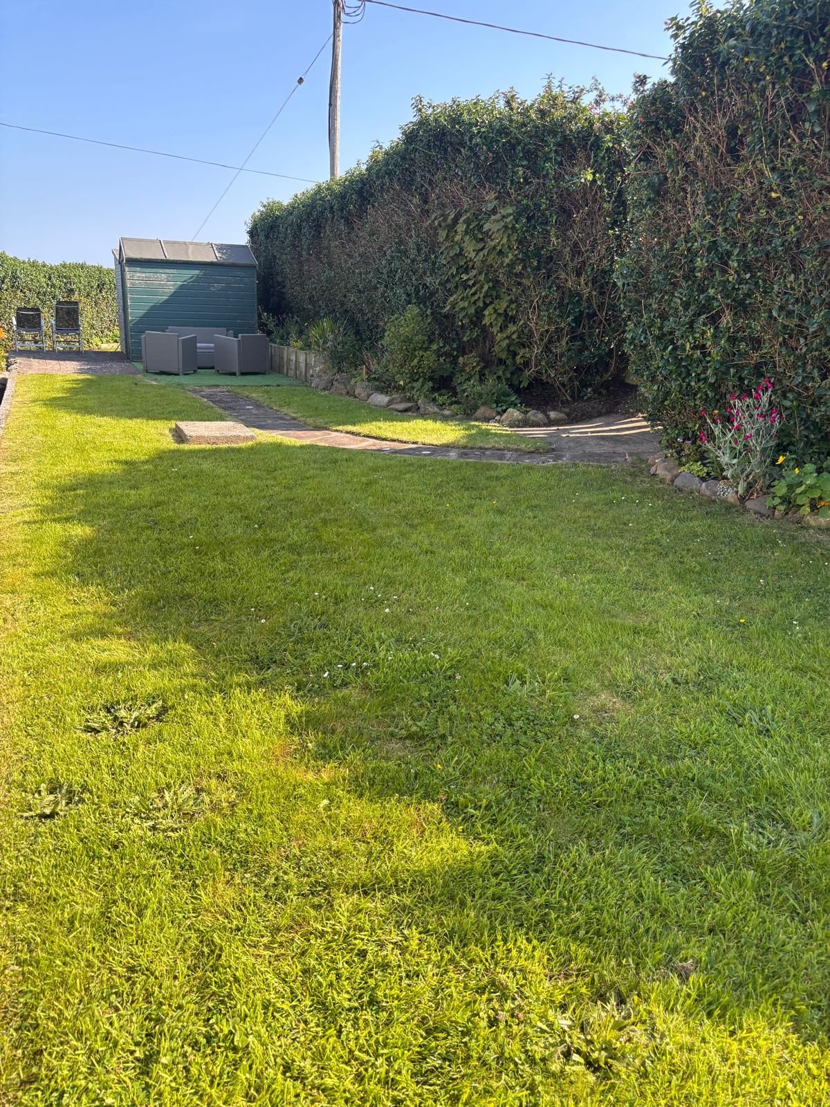Un área verde bien cuidada con un sendero pavimentado, un cobertizo verde y un seto frondoso.