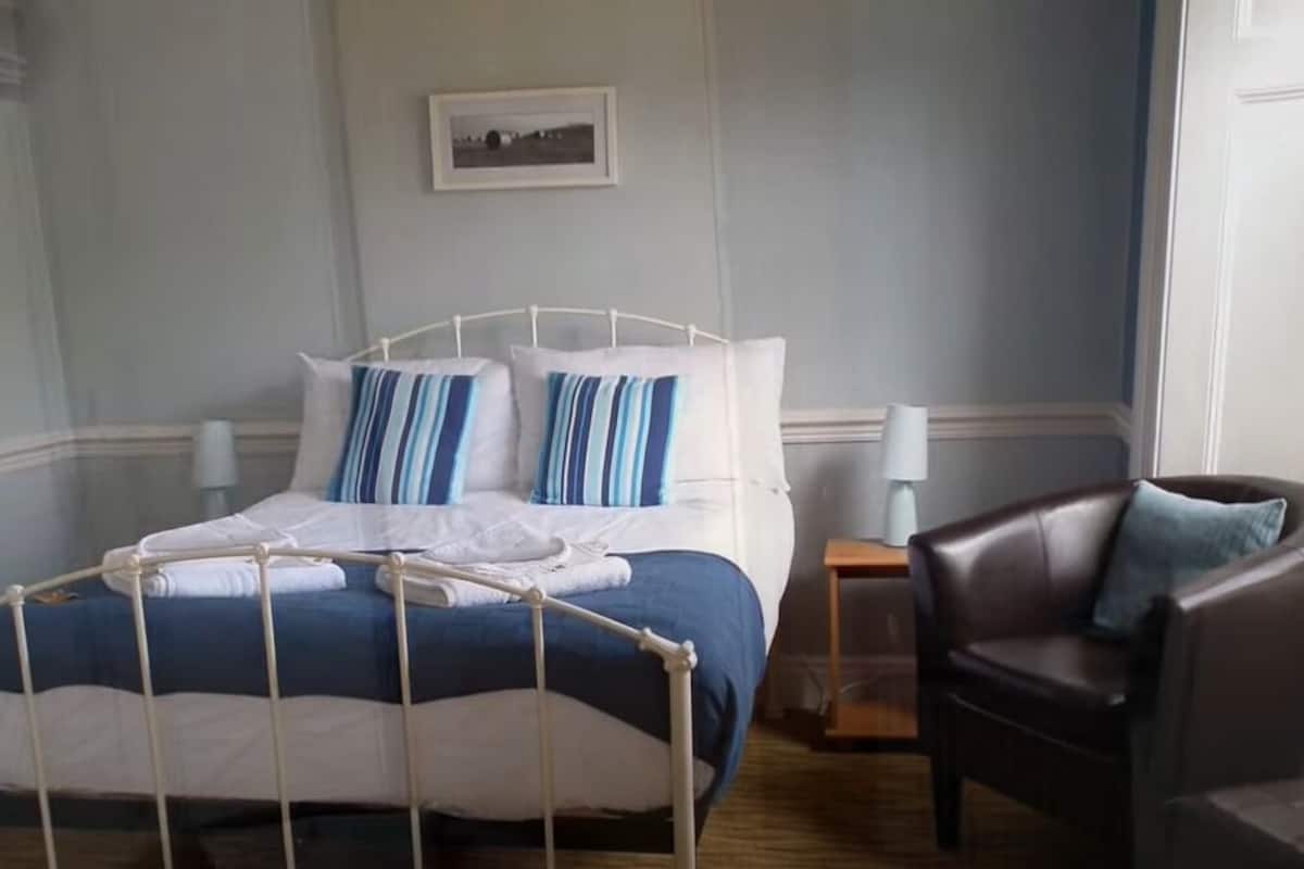 A neatly made bed with a white metal frame, a blue and white striped pillow, and a white blanket. There is a dark brown leather chair with a blue pillow to the right of the bed.