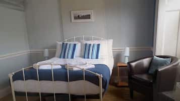 A neatly made bed with a white metal frame, a blue and white striped pillow, and a white blanket. There is a dark brown leather chair with a blue pillow to the right of the bed.