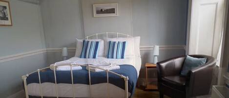 A neatly made bed with a white metal frame, a blue and white striped pillow, and a white blanket. There is a dark brown leather chair with a blue pillow to the right of the bed.