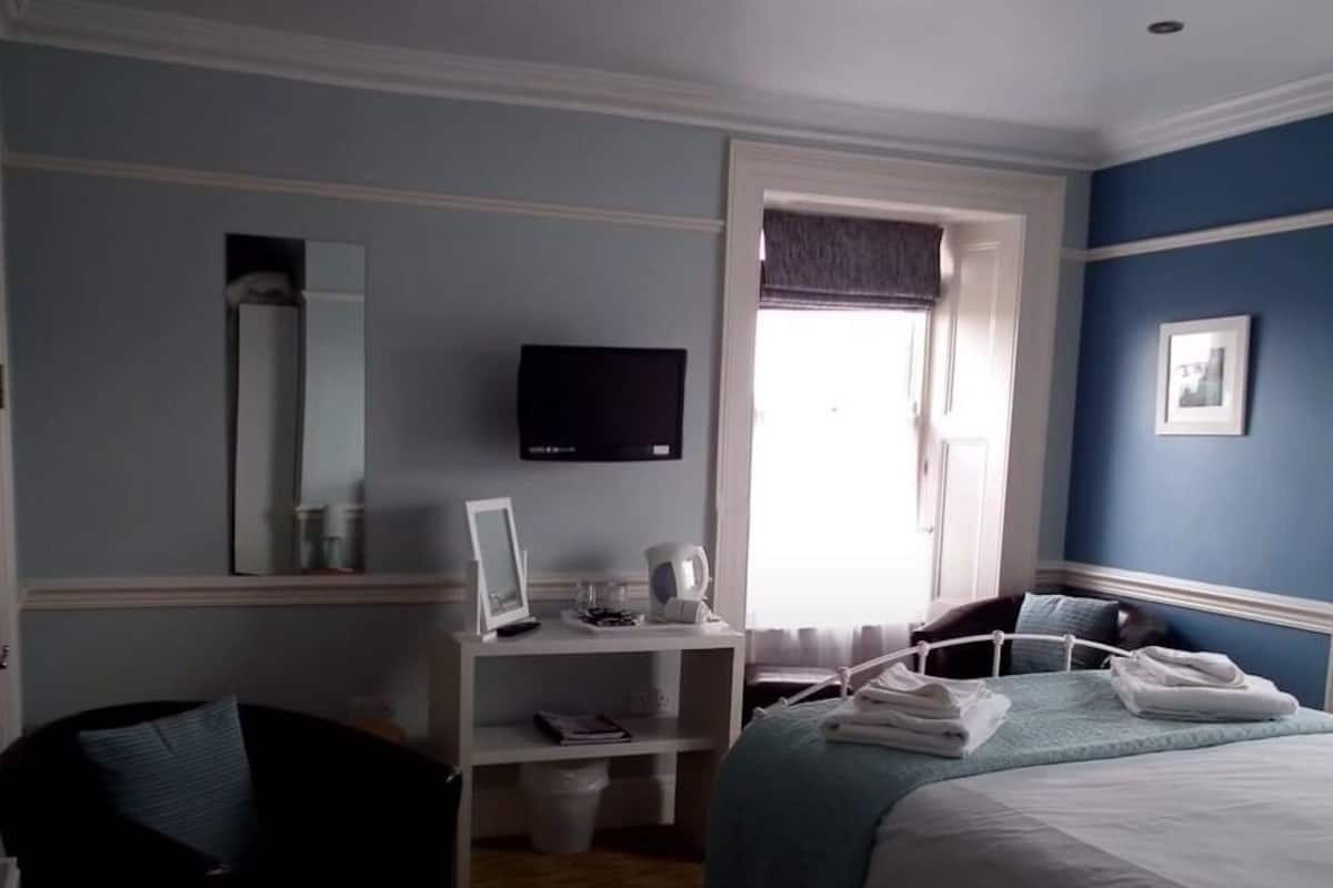 A bedroom with a bed, a desk with a computer, a television, a window with curtains, and a framed picture on the wall.