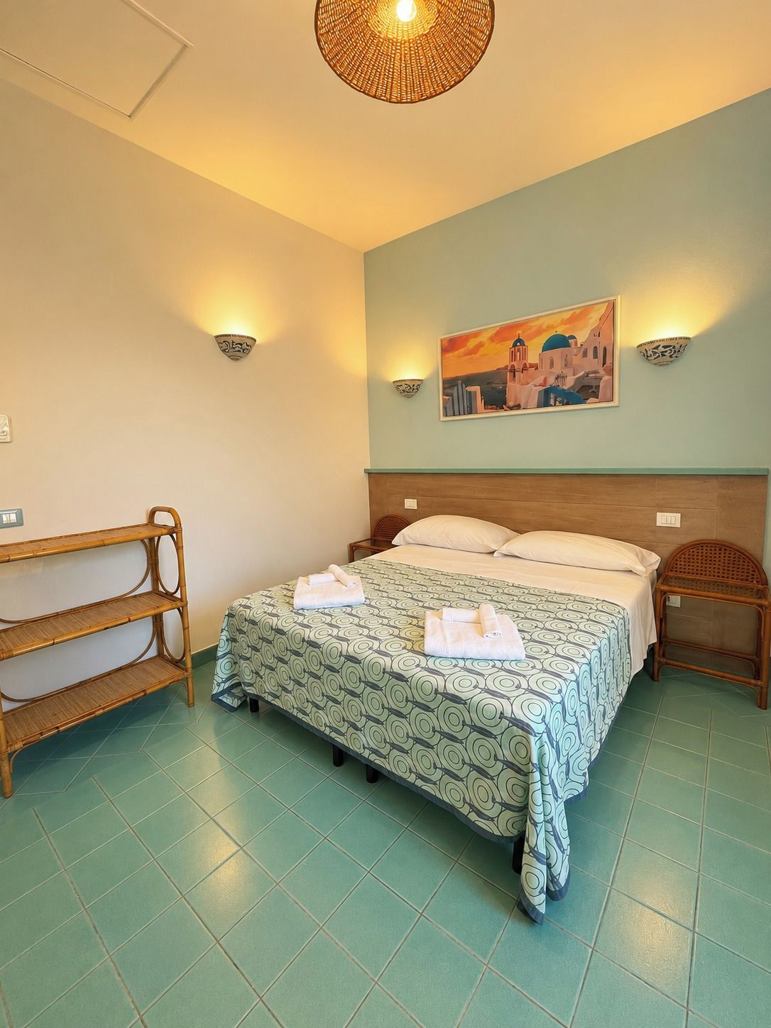 A hotel room with a bed, two wall-mounted lamps, a painting, a wooden shelf, and a rattan chair.