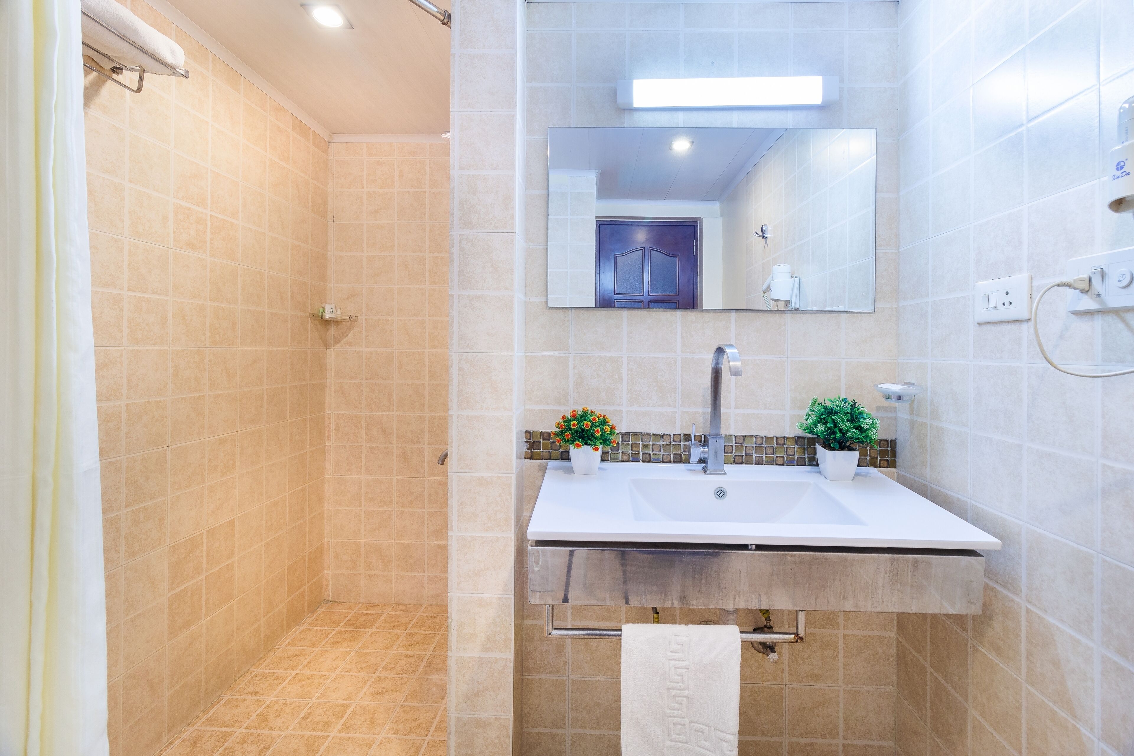 Un baño moderno con baldosas color beige, un lavamanos blanco, un espejo y dos plantas en macetas.