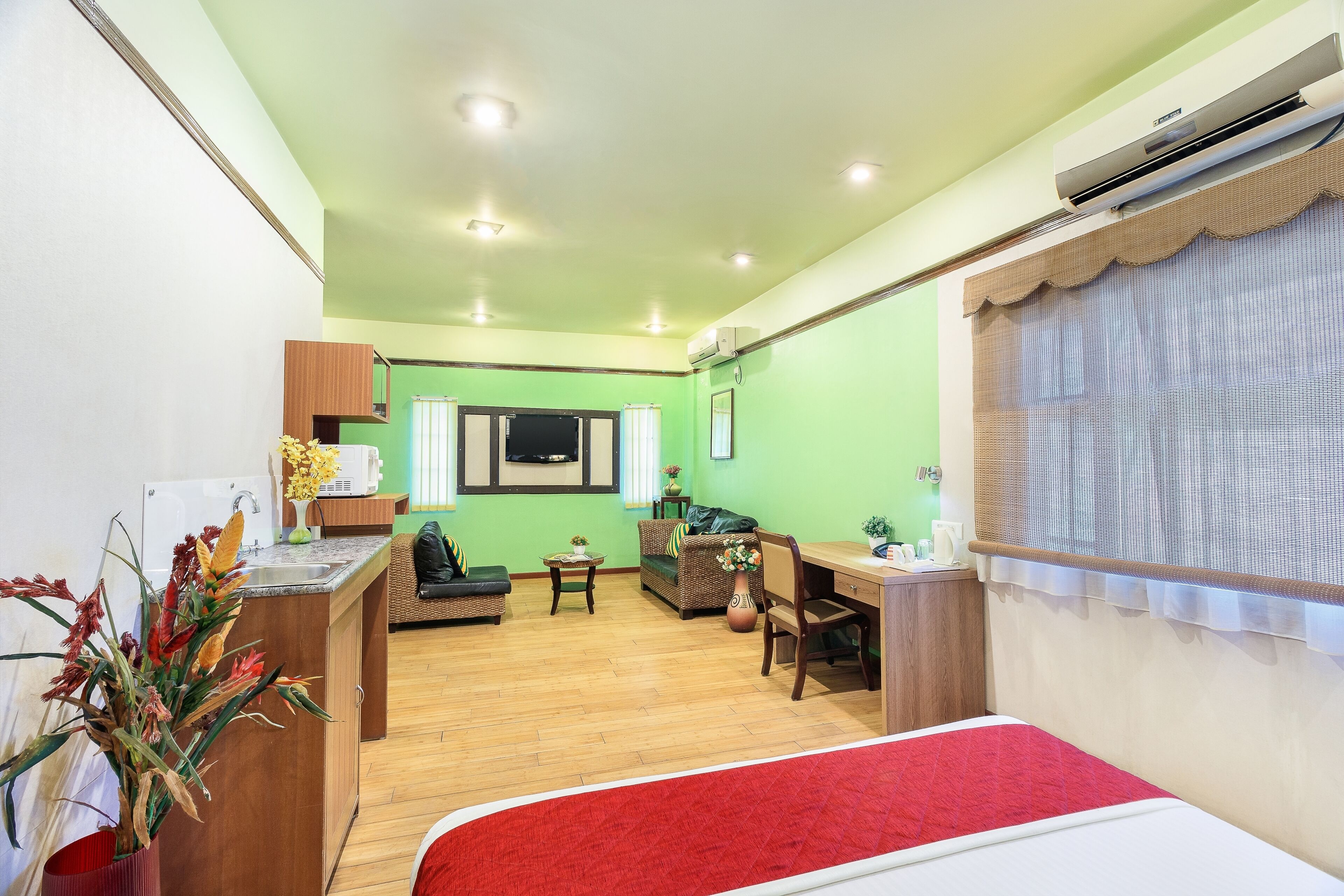 A room with a bed, a desk, a chair, a sofa, a small table, a plant, and an air conditioning unit.
