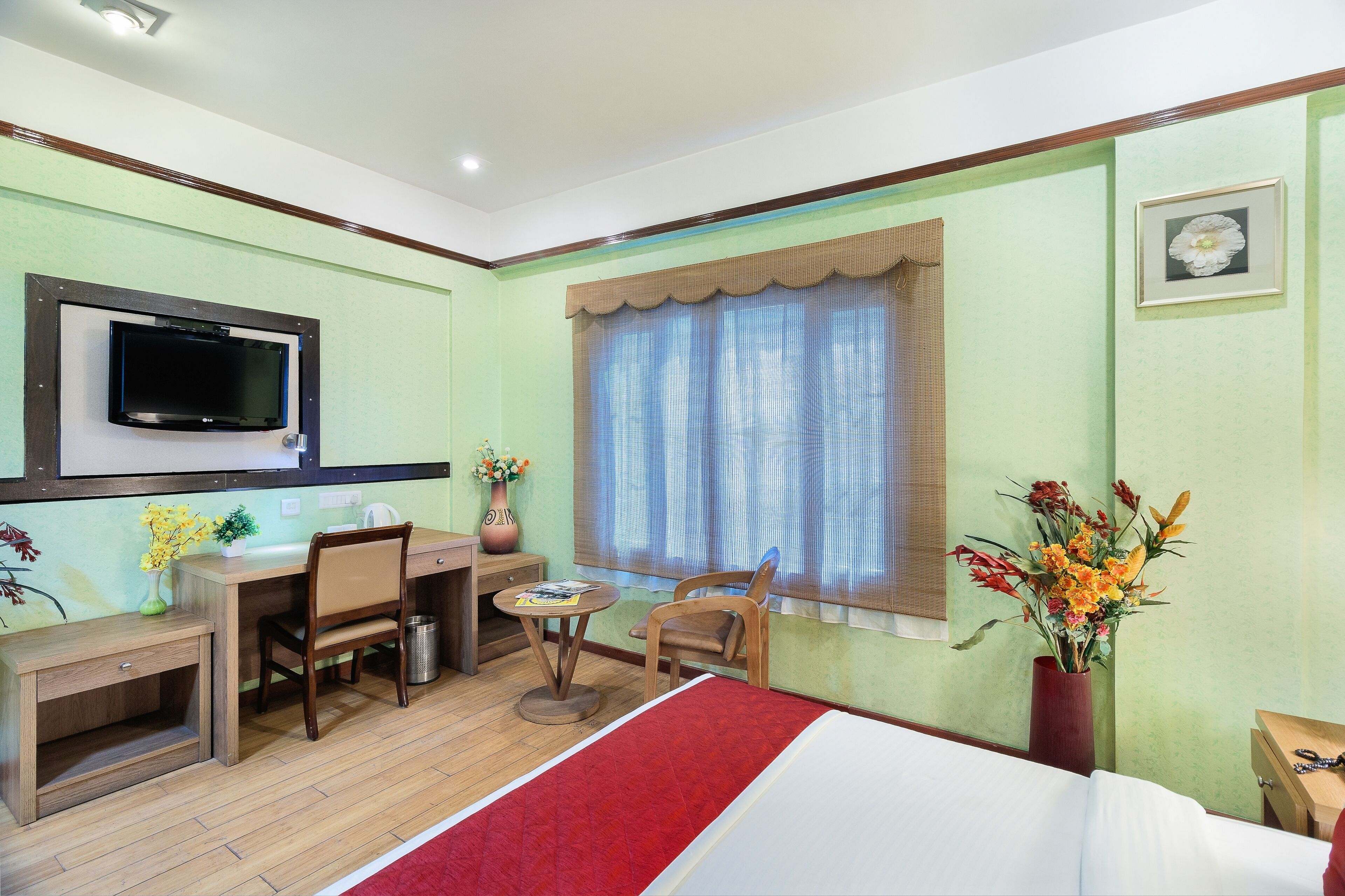 Une chambre d’hôtel comprenant un lit, un bureau, une chaise, une télévision, une fenêtre avec des rideaux et un vase avec des fleurs.