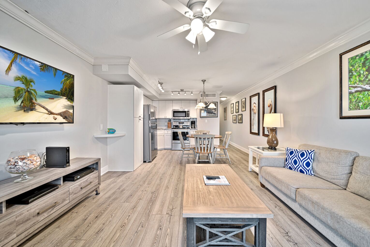 A modern living room with a wooden floor, a ceiling fan, a flat-screen TV, a dining area with a table and chairs, and a kitchen in the background.