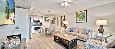 A spacious living room with a wooden floor, a sofa, a coffee table, and a dining area with chairs. There is a kitchen in the background with modern appliances.