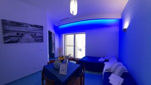 Une pièce comprenant une table à manger, des chaises, un canapé et un lit. Il y a une fenêtre avec des stores, un luminaire au plafond et un tableau accroché au mur.