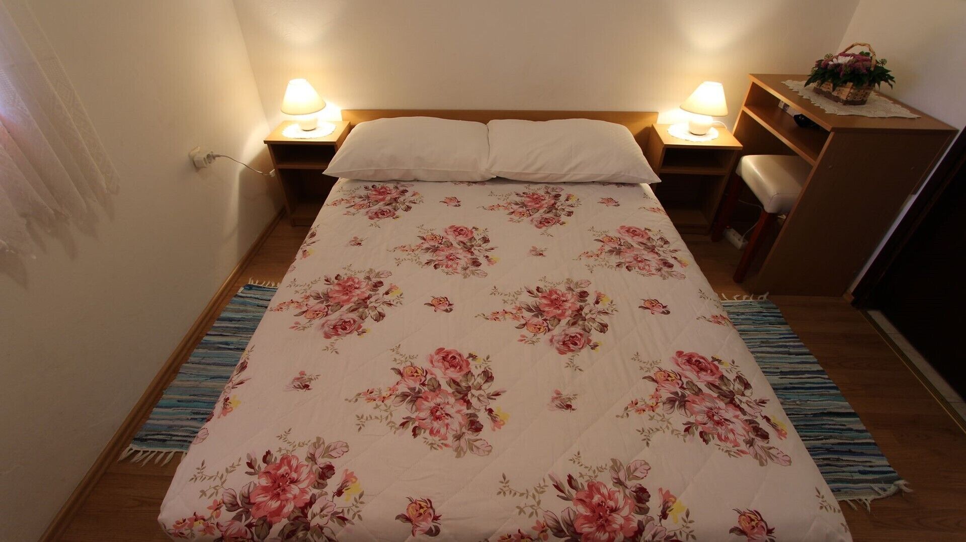 Un dormitorio con colcha estampada, dos lámparas de noche, una mesita de madera y una pequeña mesa con una cesta.