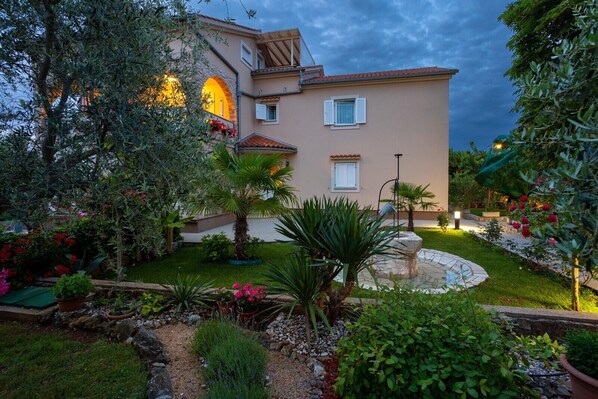 A well-maintained garden with a variety of plants and a fountain, surrounded by a building with a tiled roof and a stone pathway leading to the entrance. - Attractive apartment Janeš Studio for 3 persons on the island of Krk (Sveti Anton)